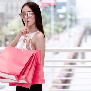 Beautiful asian woman holding red shopping bags posing hush quiet sign for special private sale with busy shopping district and roads in background. She spent all her husband money.